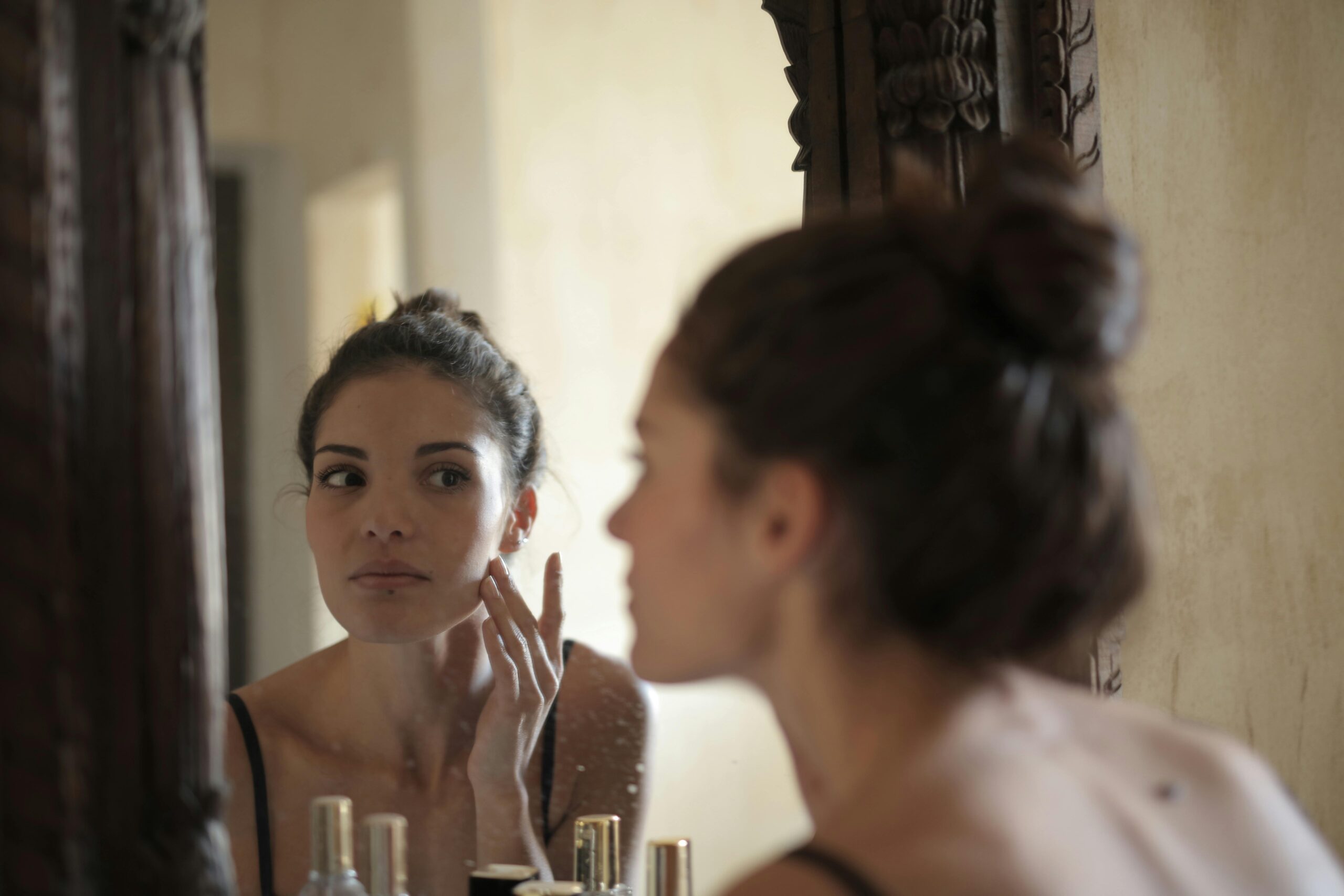 A woman checks her skin in the mirror, engaging in her morning skincare routine indoors.