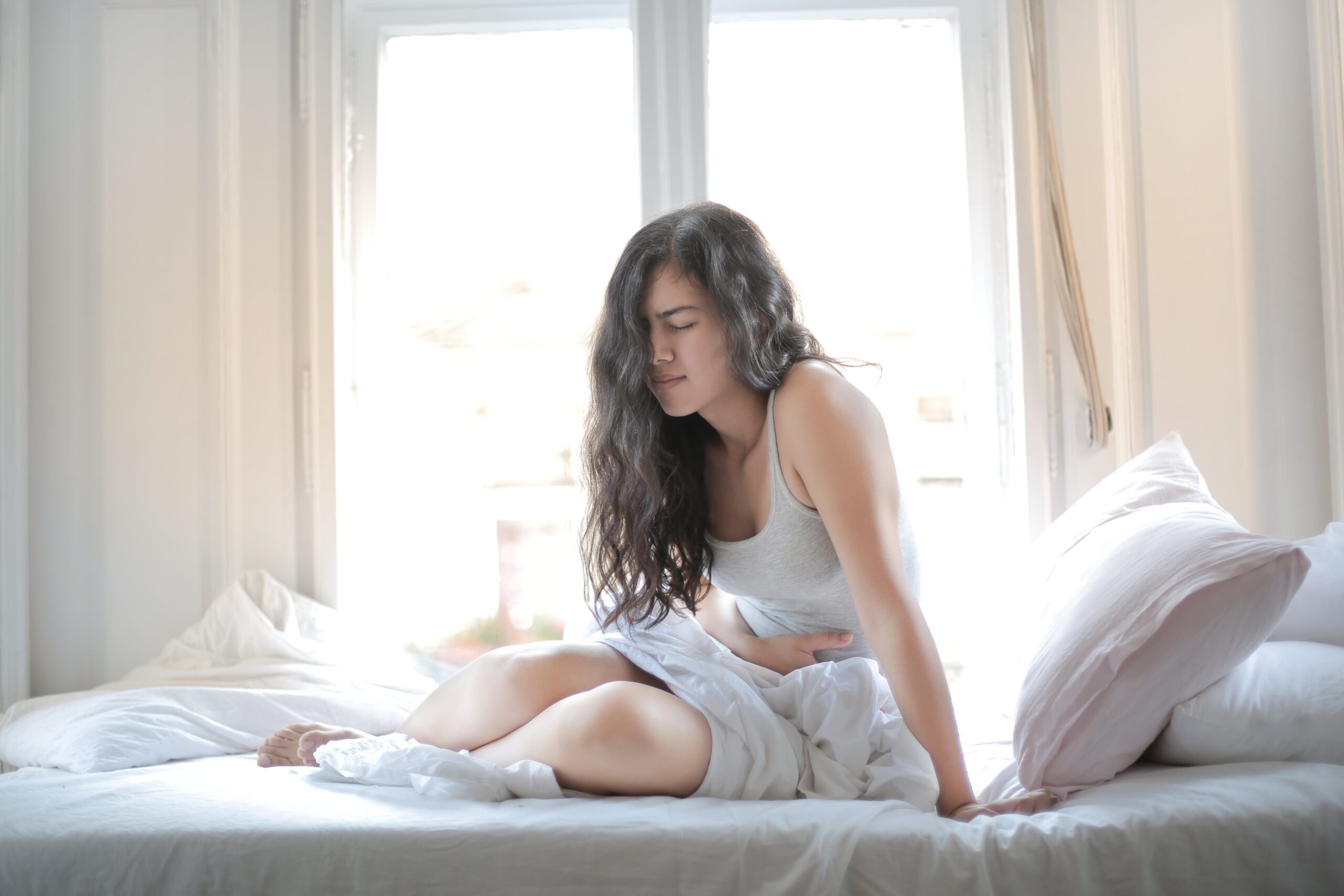 Woman sitting on bed holding her stomach, showing signs of discomfort or pain indoors.