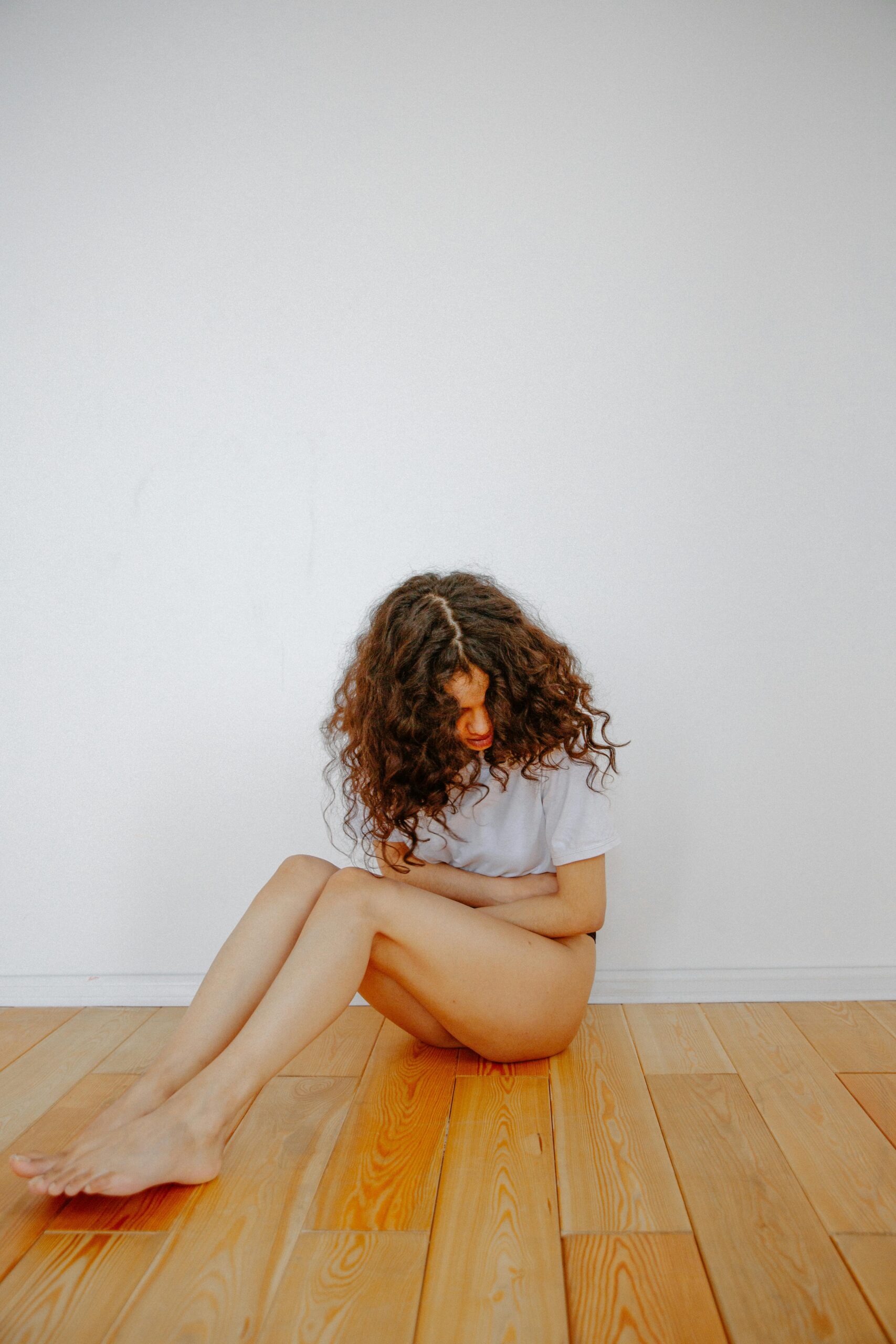 A woman sits on a wooden floor clutching her stomach in discomfort.