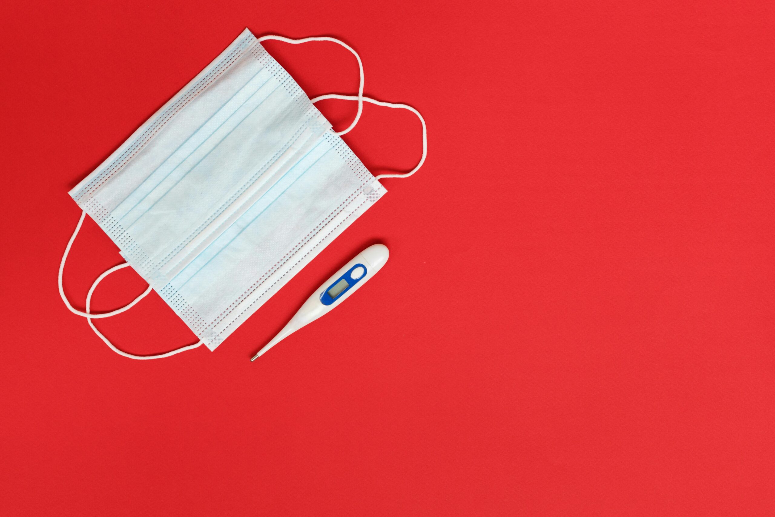 Flat lay of a surgical mask and digital thermometer on a red background, symbolizing health and safety.