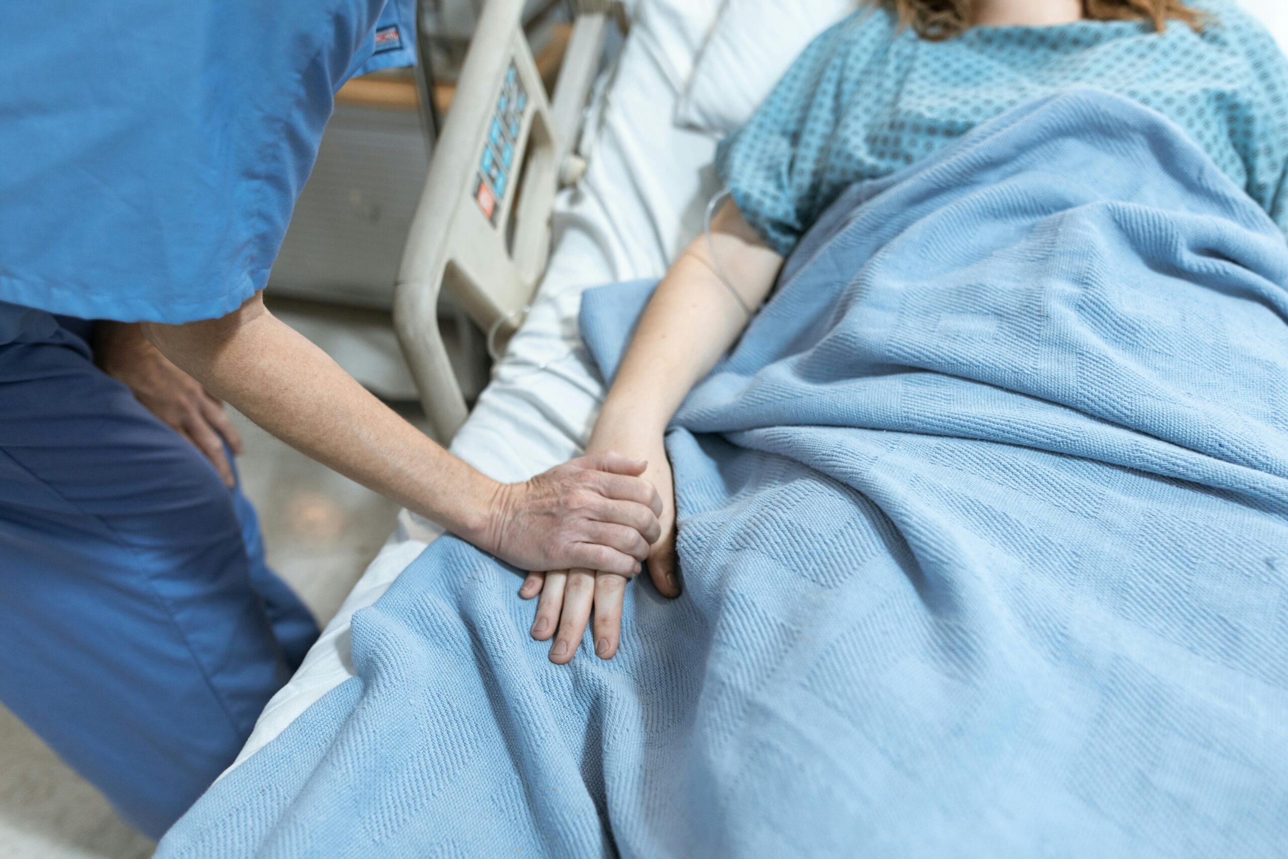 A nurse in scrubs offers comfort by holding a patient's hand in a hospital bed.