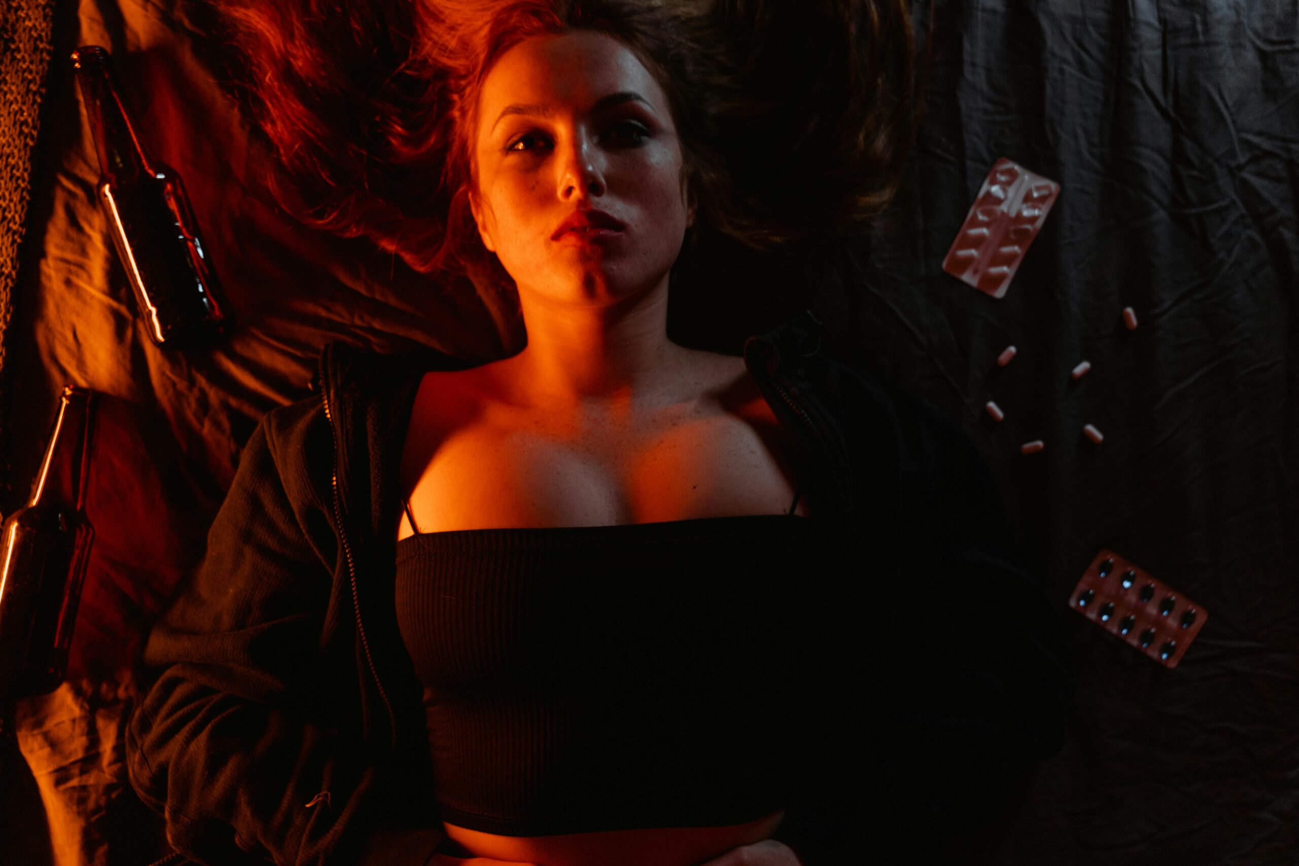 A woman lying on a bed surrounded by bottles and pills, illuminated by moody lighting.