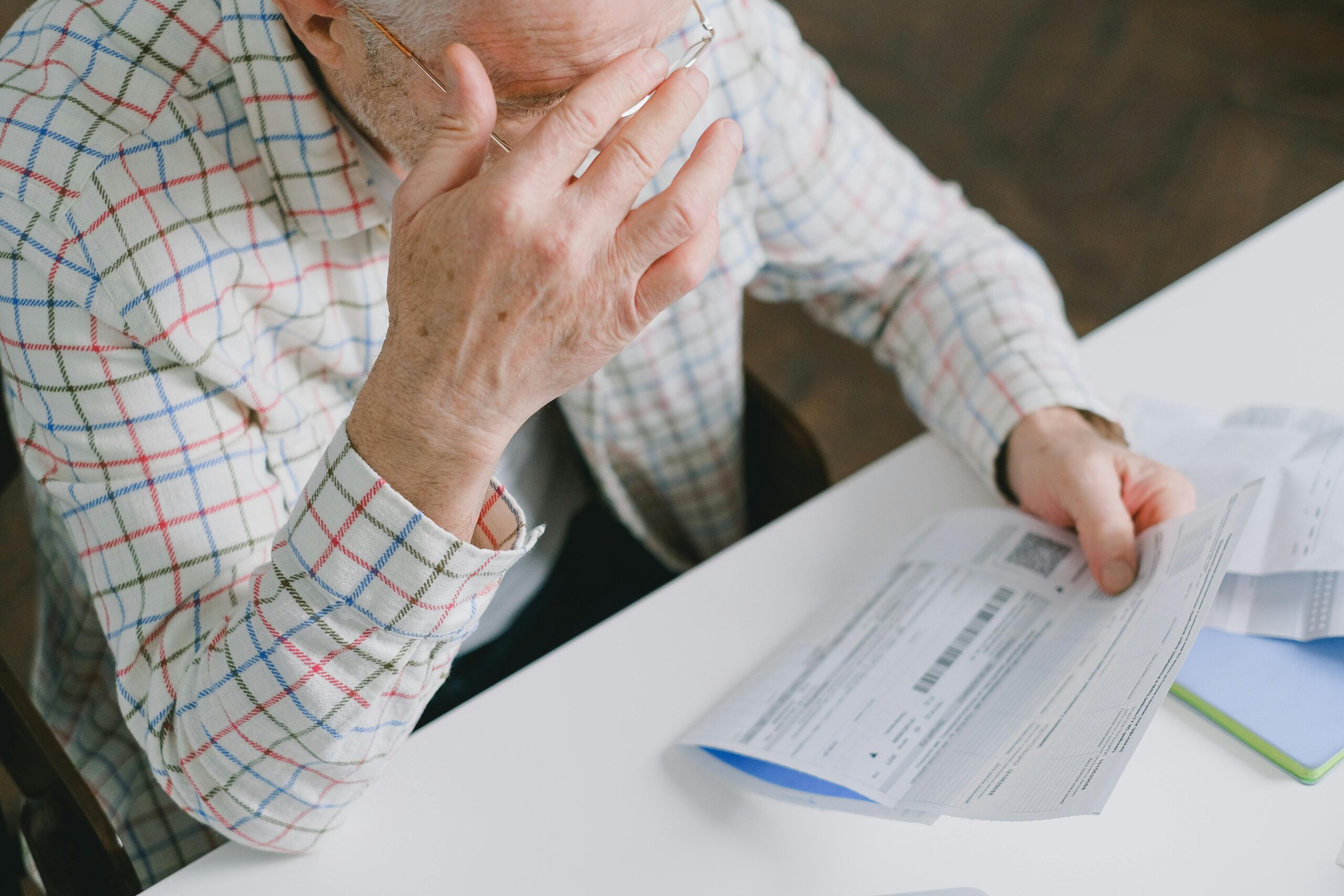 Elderly man in patterned shirt reading and holding bills at a home table, appearing focused.