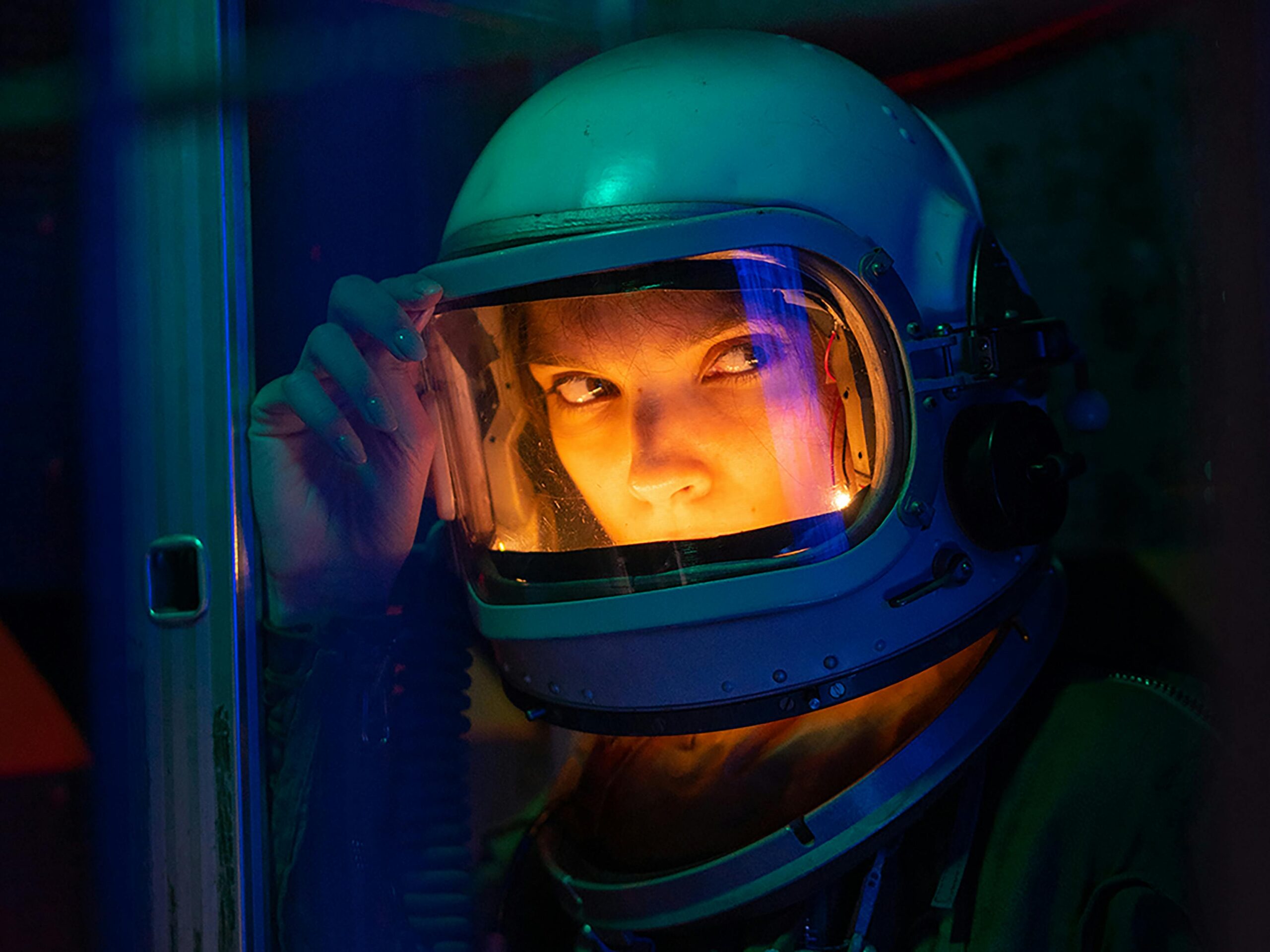 Portrait of a spacewoman in a helmet gazing intensely with colorful lighting.