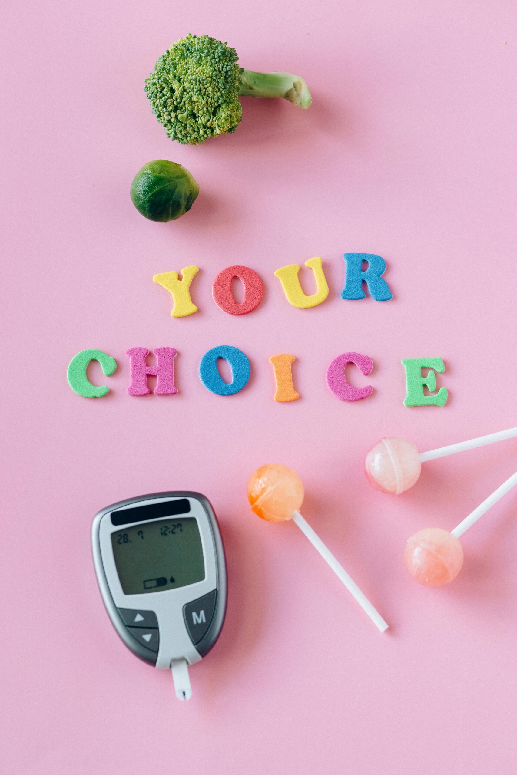 Flat lay of glucometer, vegetables, candies, and text 'Your Choice' on pink background.