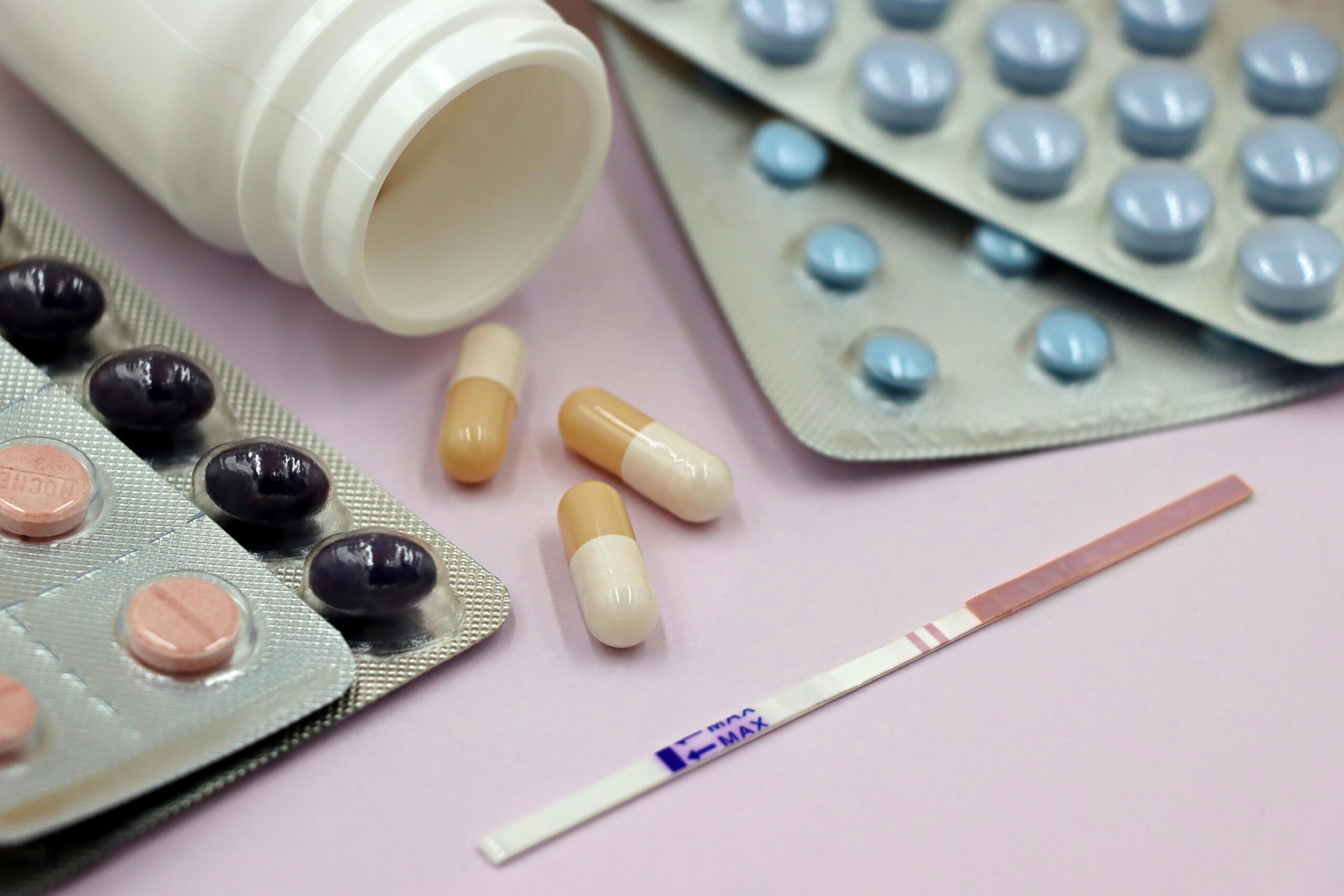 Image featuring a pregnancy test strip surrounded by various pills and capsules on a pastel background.