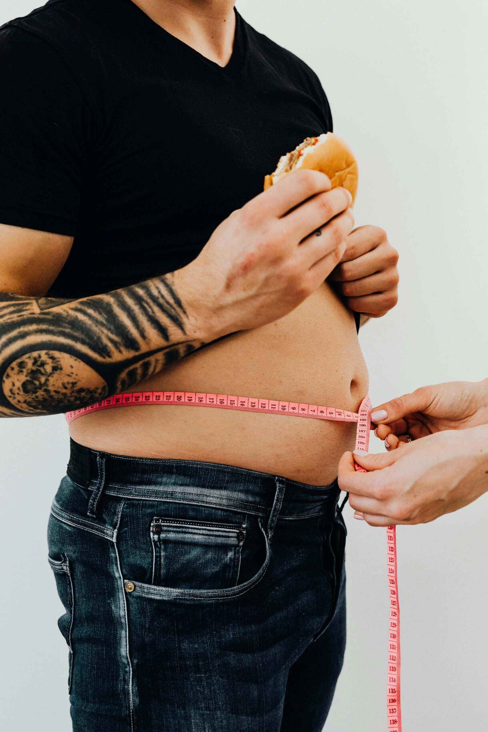 Close-up of a man holding a burger while measuring his waistline with a tape, highlighting unhealthy eating habits.