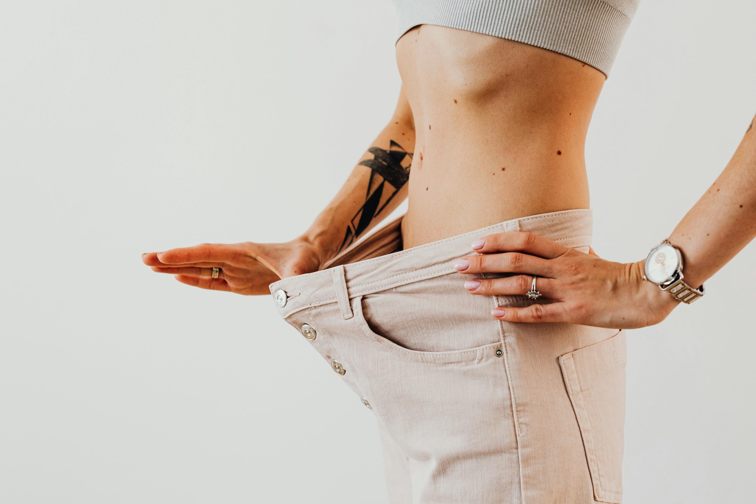 Close-up of a woman in oversized pants showcasing weight loss progress. Studio shot with neutral background.