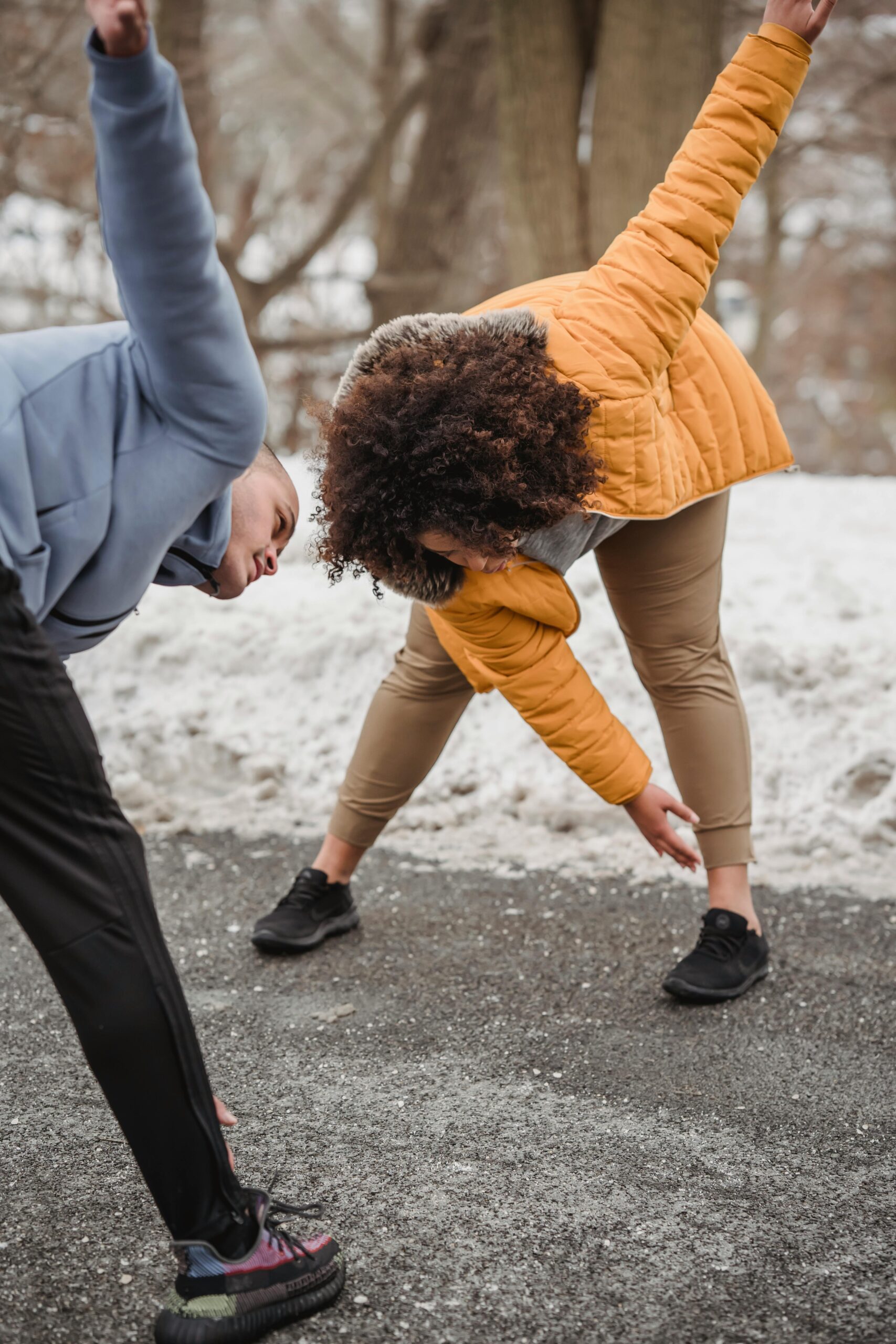 Active plum plus size African American woman with black personal instructor bending forward while doing stretching exercise on walkway during training in winter time