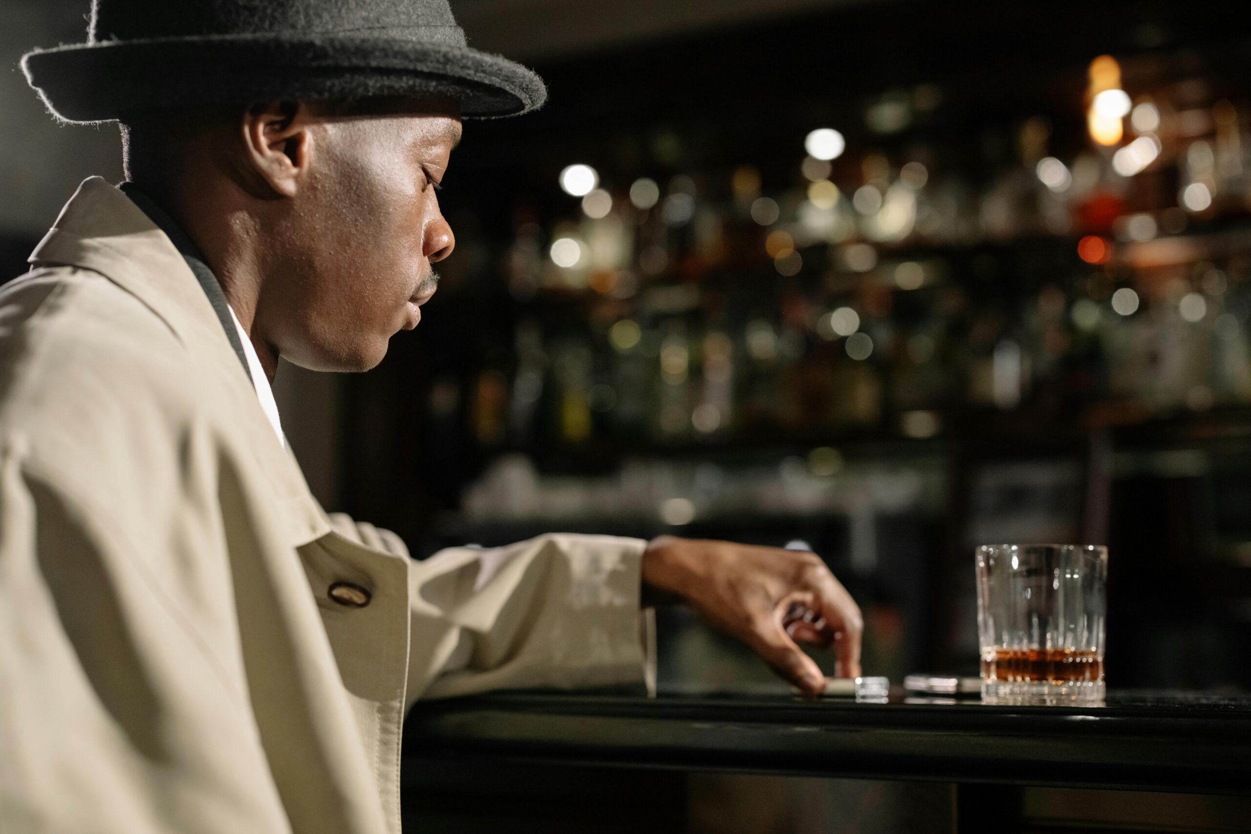 Side view of a thoughtful man in a hat, sitting at a bar with a whiskey glass in a moody atmosphere.