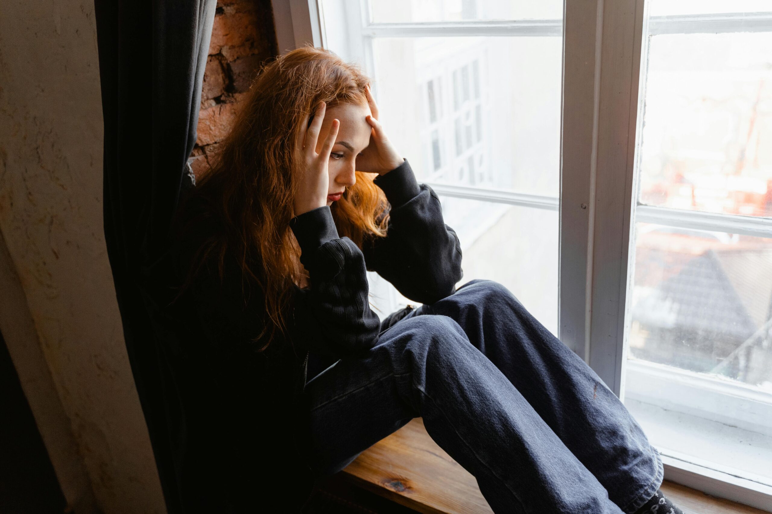 Woman sitting by a window, deep in thought, conveying emotions like distress and anxiety.