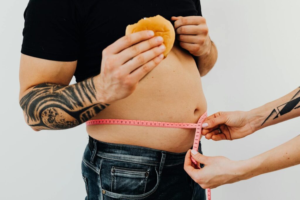 A man with tattoos holding a burger as another person measures his waistline with a tape measure.