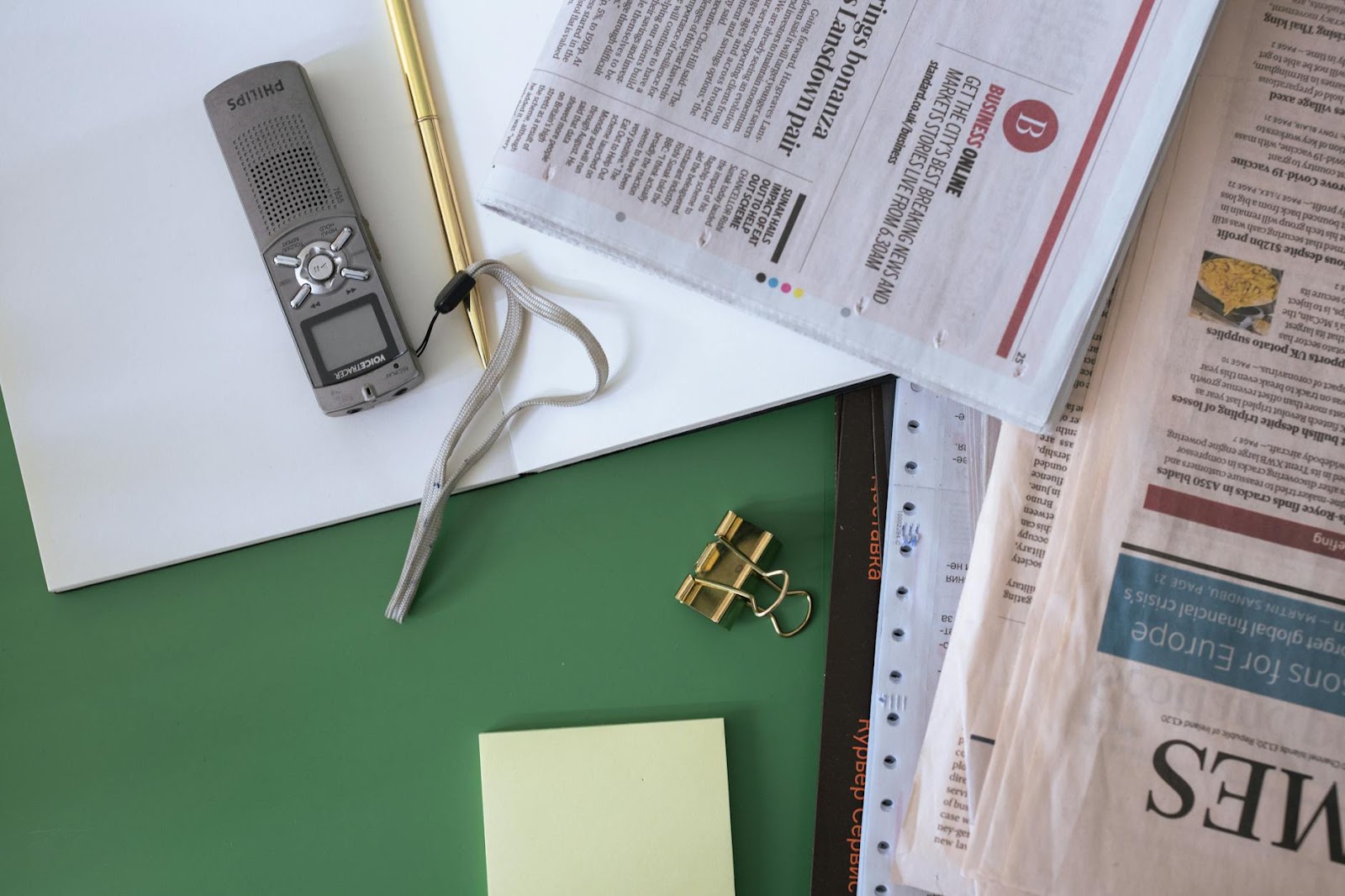 Flat lay of an office desk with newspapers, notepad, and recorder on a green surface.