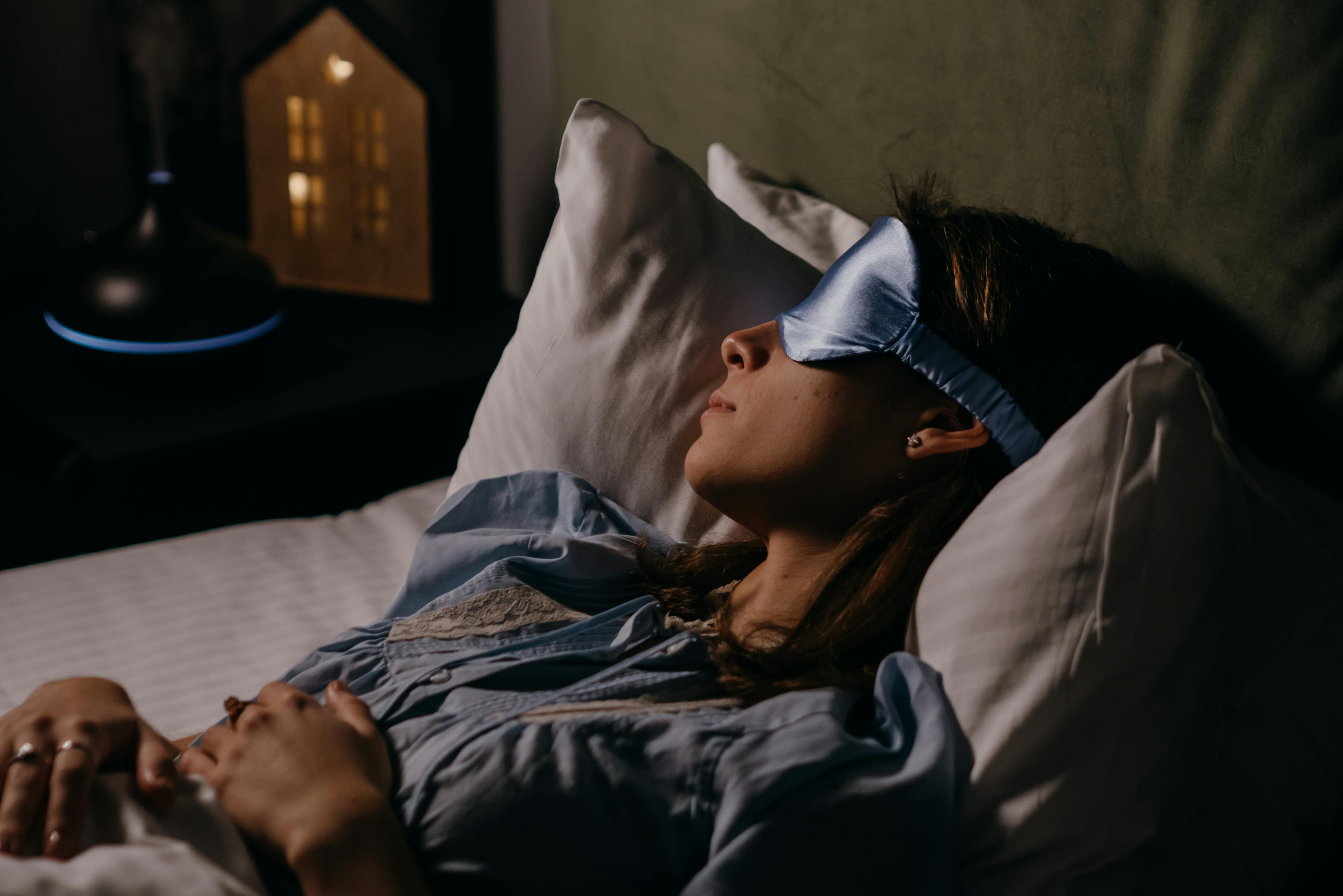 A woman sleeping peacefully wearing a blue eye mask in a cozy, dimly lit bedroom.